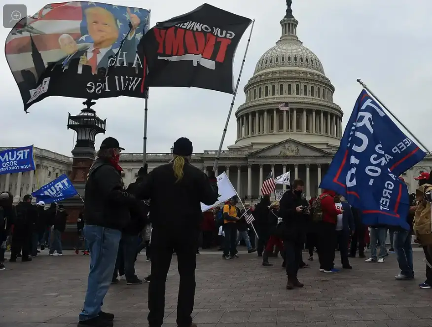 A crowd of people stands in front of the U.S. Capitol building, holding various flags and banners. Prominently visible are…