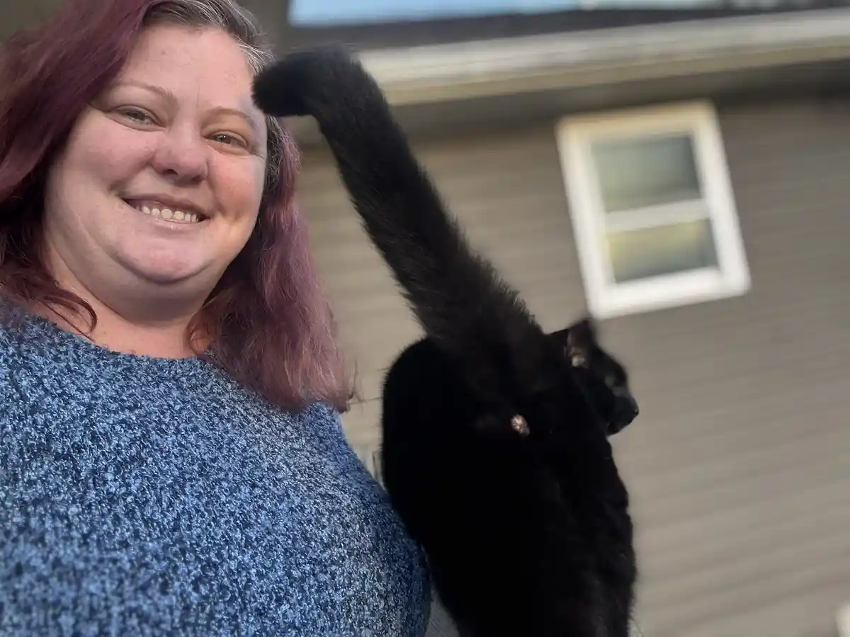 A woman with reddish-purple hair and a blue sweater smiles at the camera while a black cat stands beside her, its tail…