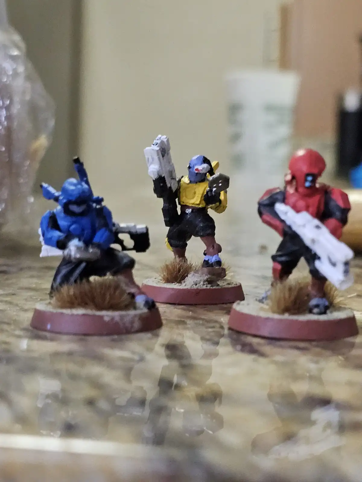 Three painted miniature figurines, resembling sci-fi or fantasy soldiers, stand on bases with grass tufts on a reflective…