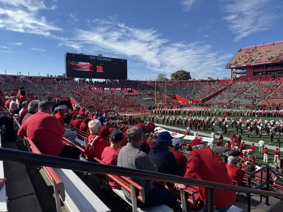 A wide shot of a crowded football stadium, likely Rutgers, with fans in red attire filling the stands under a blue sky. A…
