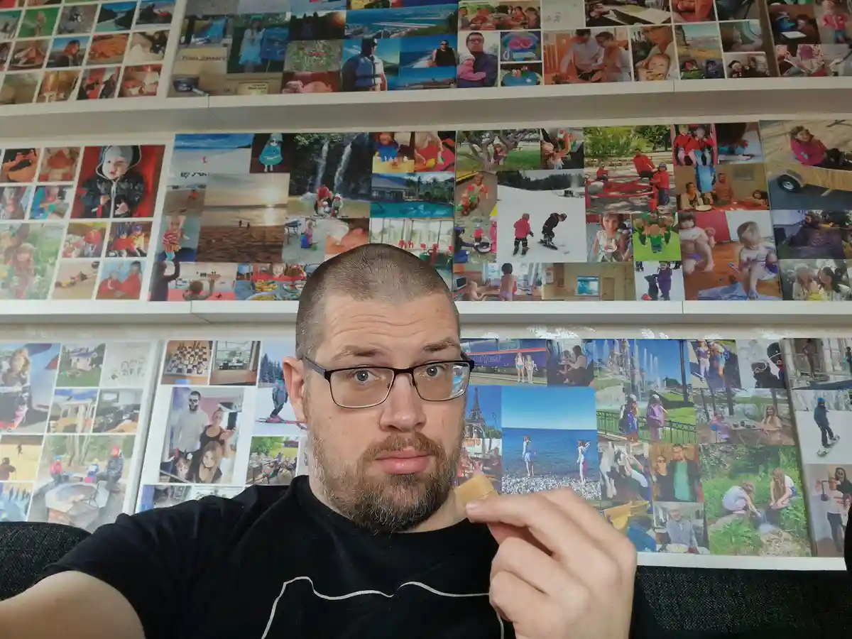 A man with glasses and a beard holds a small piece of food near his mouth while looking directly at the camera with a weary…