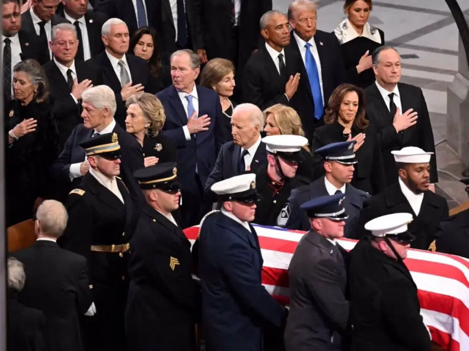 A solemn photo of U.S. presidents and their families standing in a line at a funeral, with many placing their hands over…
