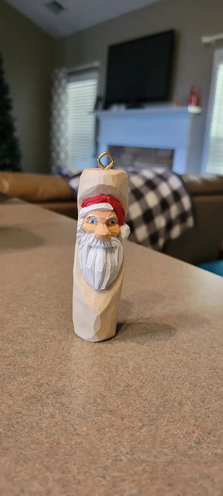 A hand-carved wooden Santa Claus ornament with a red hat and white beard sits on a countertop, featuring a gold loop at the…
