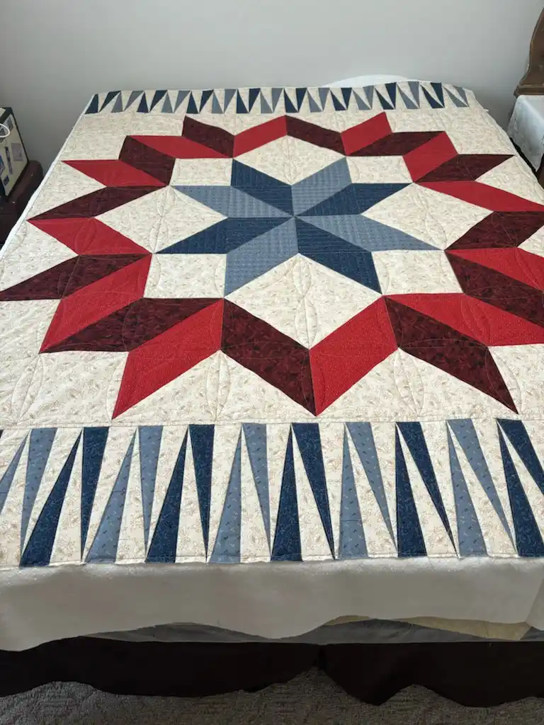 A handmade quilt with a large central star pattern made of red and blue fabric pieces on a cream background, laid over a…