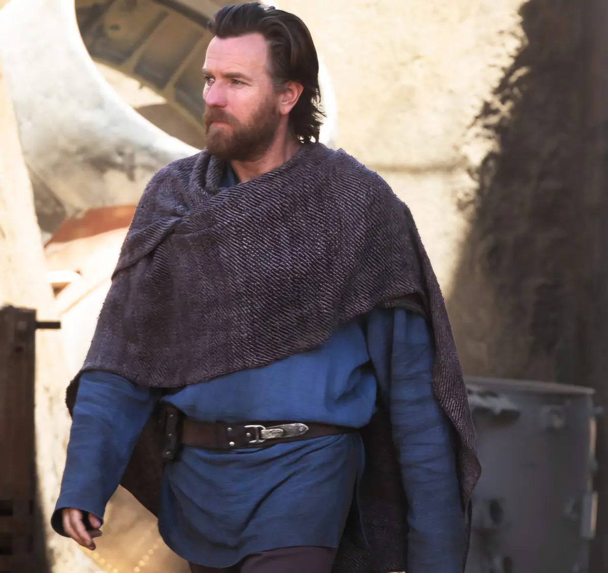A man with a beard and dark hair, wearing a blue tunic and a dark textured cloak, stands in a desert-like setting with a…