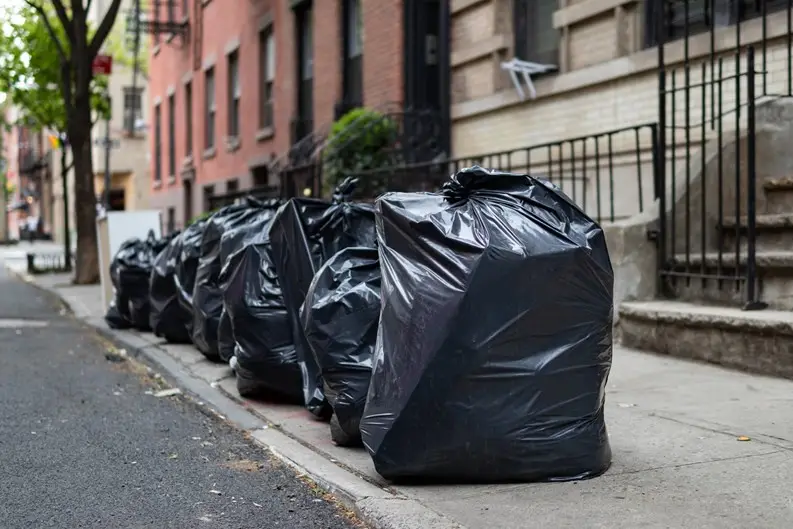 A row of large, full black garbage bags lined up along a city sidewalk next to brick buildings and fire escapes. The bags…
