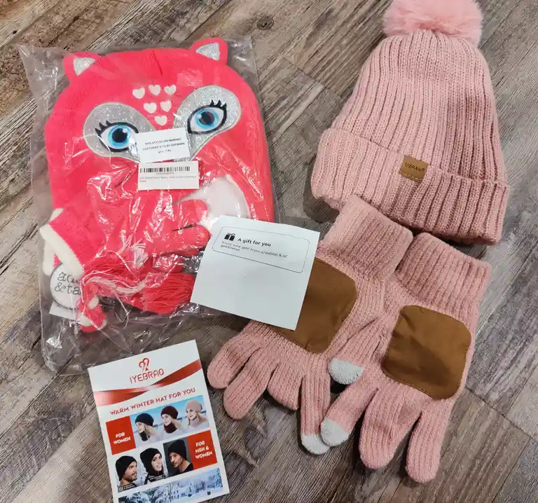 A flat-lay photo shows two winter accessory sets on a wooden floor: one is a pink fox-themed hat and gloves in plastic…
