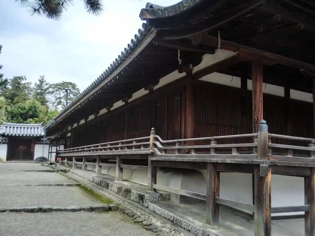 A long, traditional wooden Japanese temple building with a curved tiled roof, wooden railings, and a gravel courtyard under…