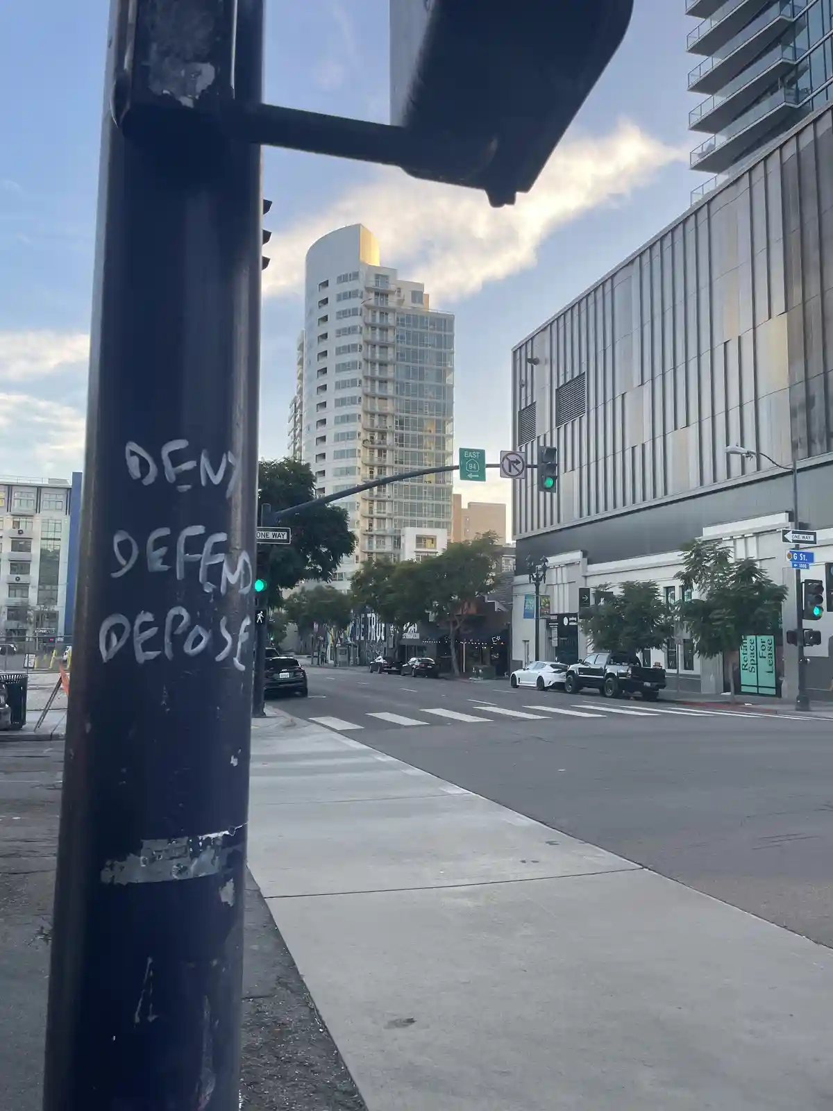 A street scene with a black pole in the foreground showing the words 'DENY', 'DEFEND', and 'DEPOSE' written in white…