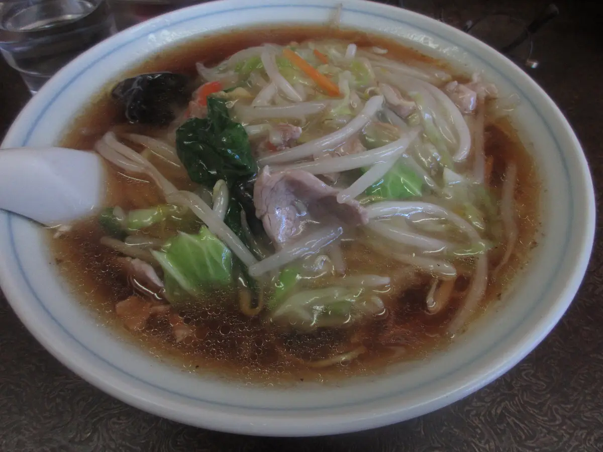 A close-up photo shows a white ceramic bowl filled with ramen soup, containing noodles, bean sprouts, cabbage, green leafy…