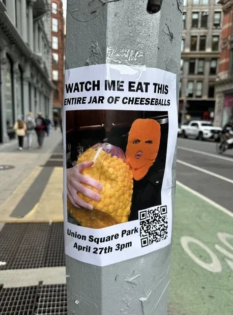 A street poster taped to a pole shows a person in an orange balaclava holding a large jar of cheeseballs, with bold text…