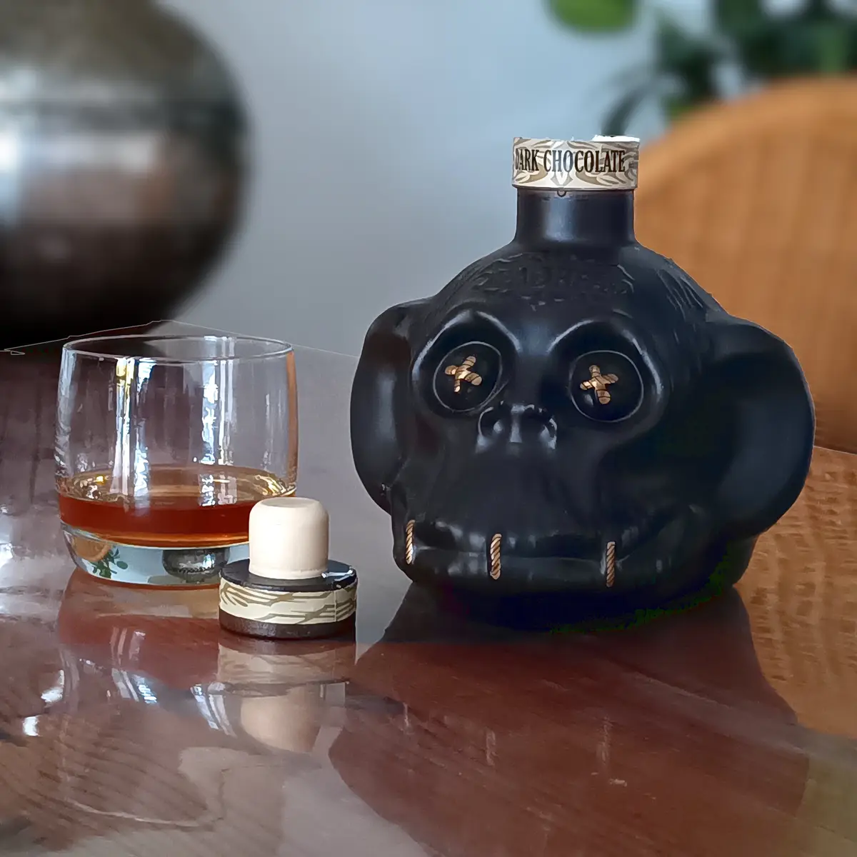 A black skull-shaped bottle labeled 'DARK CHOCOLATE' sits on a reflective table next to a glass of amber liquid and its cap…
