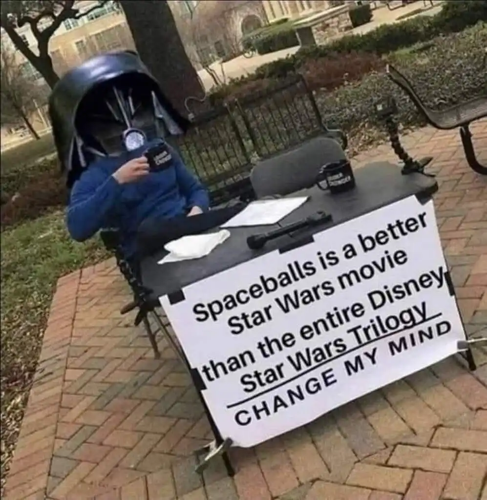 A person wearing a Darth Vader helmet sits at a small outdoor desk in a park, holding a mug, with a sign in front of them…