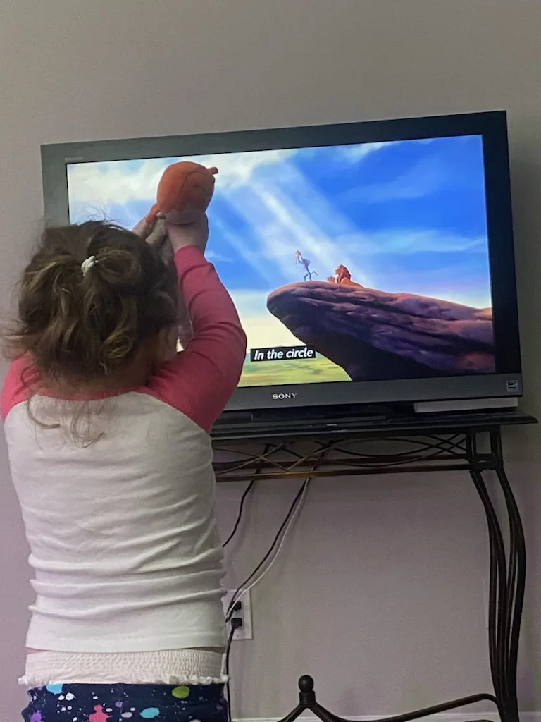 A young child, seen from behind, holds a stuffed animal up to a TV screen showing a scene from 'The Lion King' where Simba…
