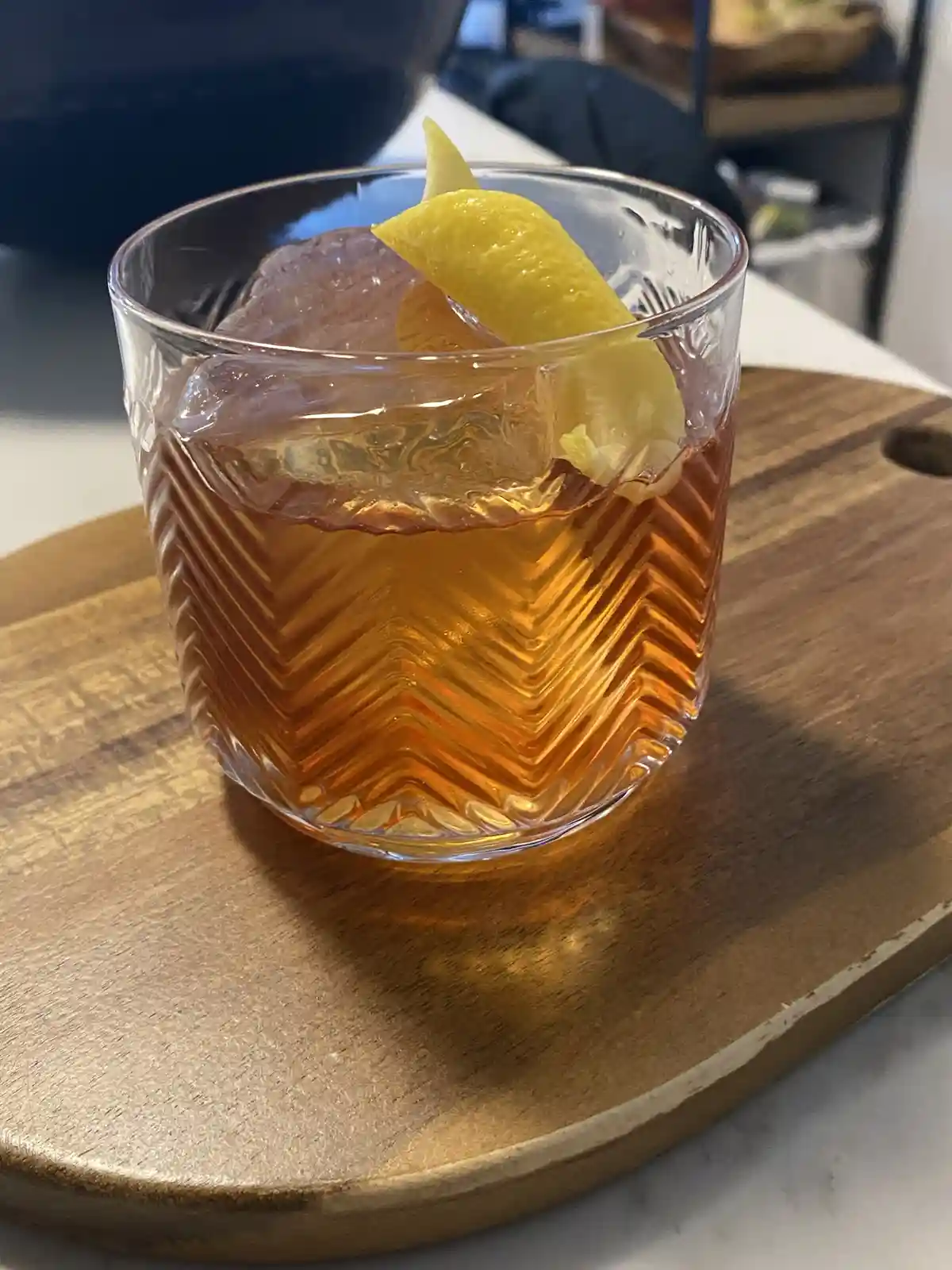 A close-up photo shows a cocktail in a textured glass with a large ice cube and a lemon wedge garnish, resting on a wooden…
