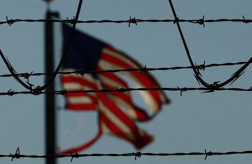 A blurry American flag waves in the background behind several strands of sharp barbed wire in the foreground. The flag's red…