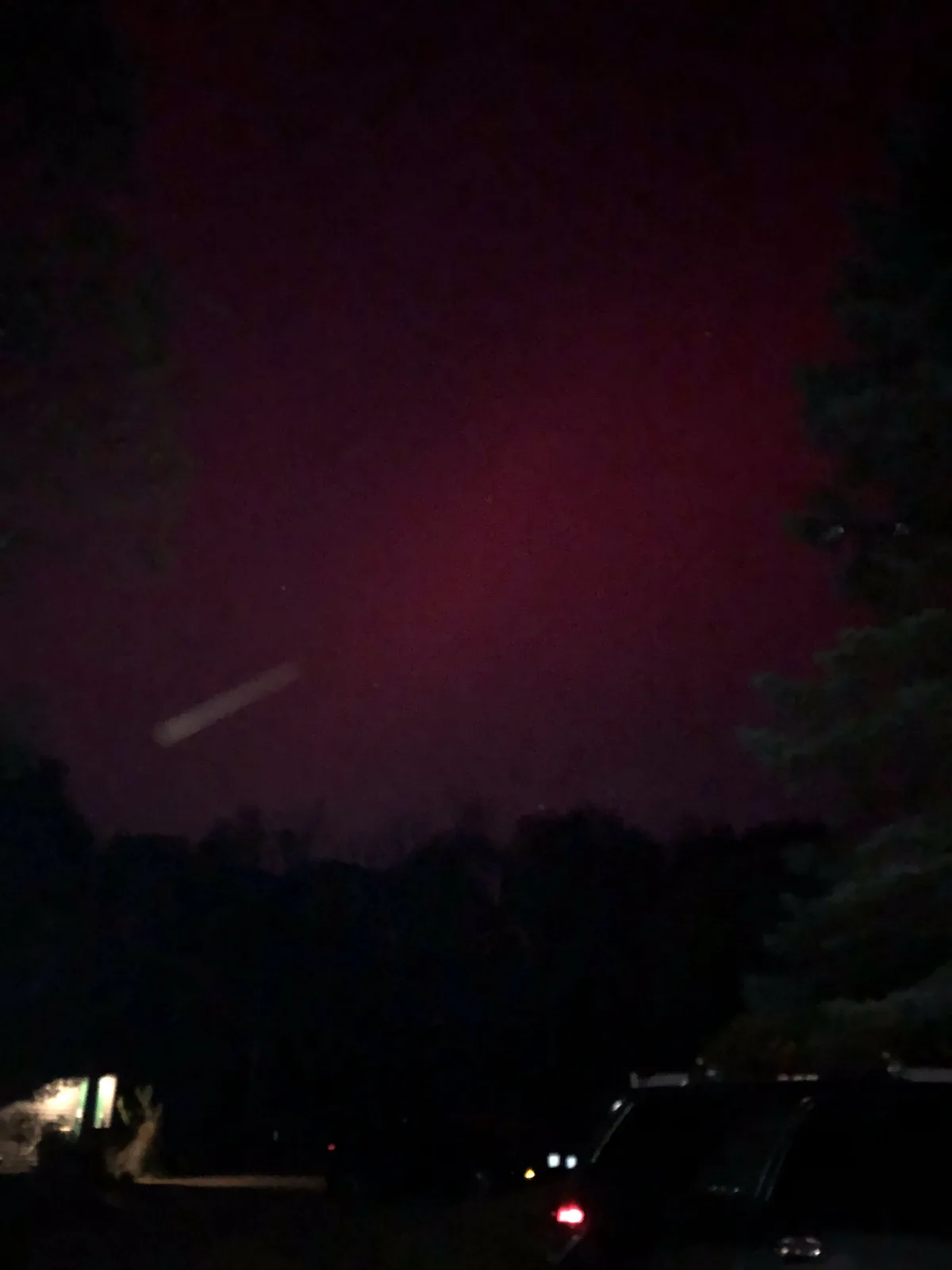 A blurry night photo shows a faint, streaking light across a dark, reddish-purple sky above silhouetted trees and a car's…