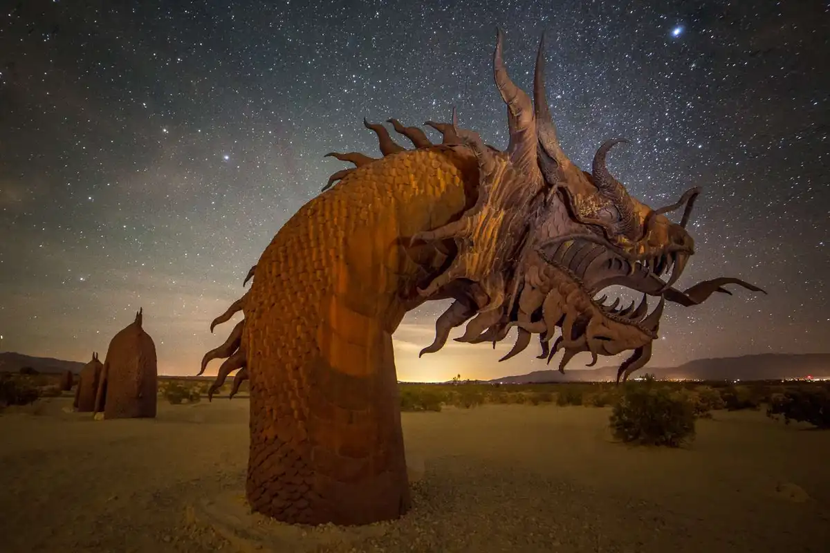 A large, intricately detailed dragon sculpture made of metal stands in a desert under a starry night sky, its head raised…