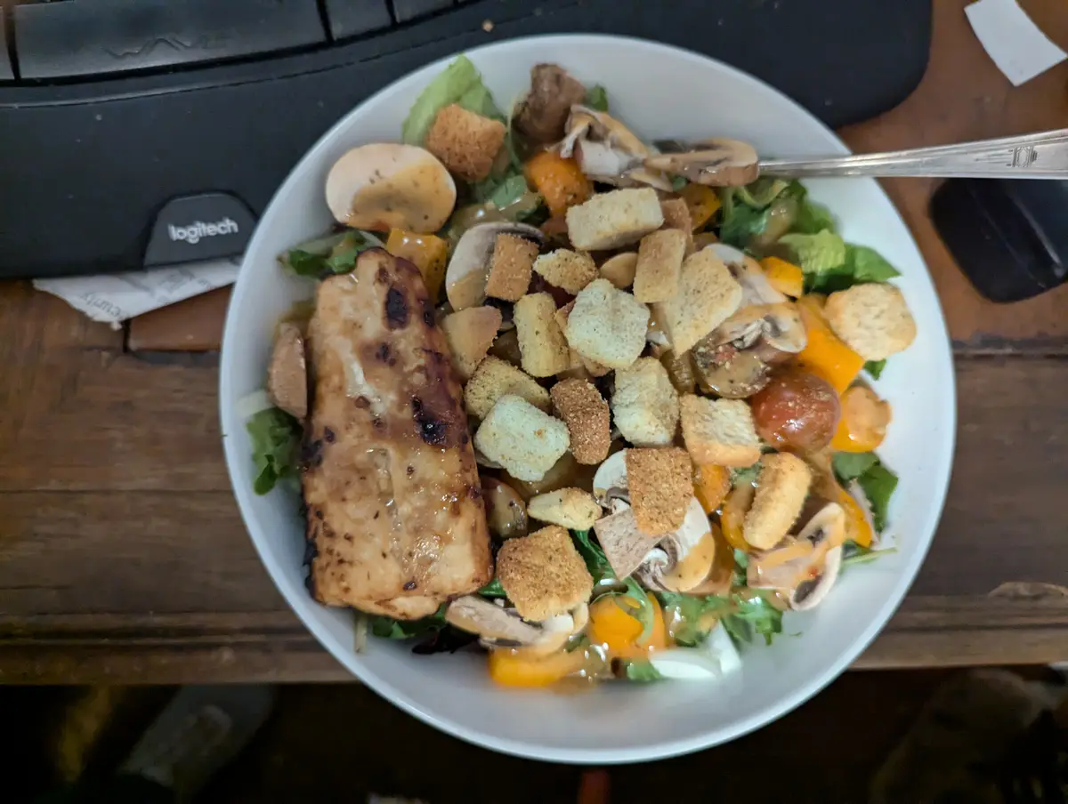 A white bowl filled with a salad containing grilled chicken, croutons, mushrooms, cherry tomatoes, and yellow bell peppers…