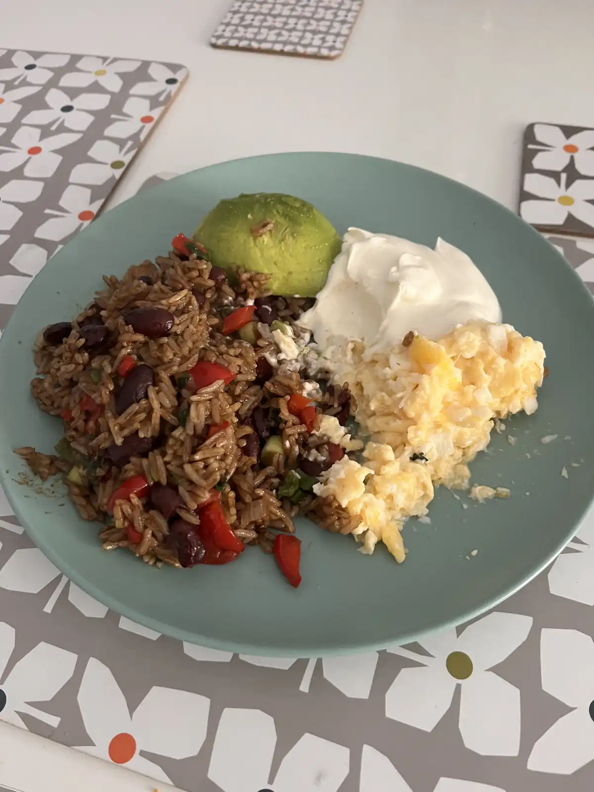 A teal plate holds a meal of brown rice mixed with black beans and red peppers, topped with a half avocado and a dollop of…
