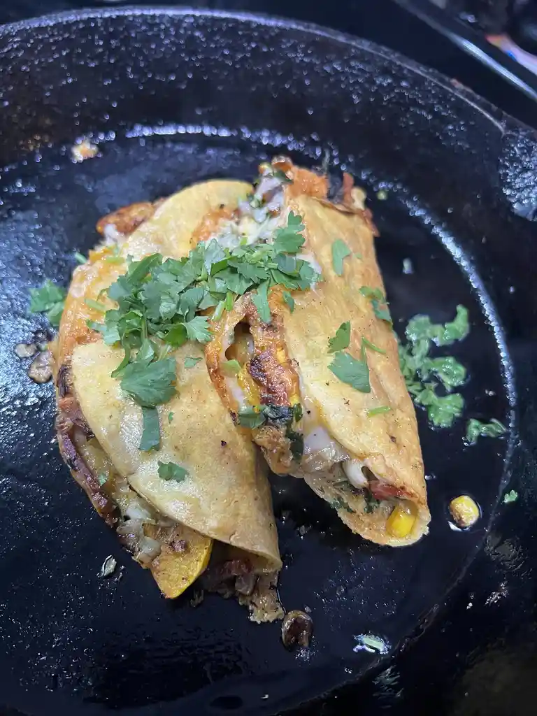 A close-up photo shows two folded, golden-brown tortillas filled with melted cheese, corn, and other ingredients, garnished…