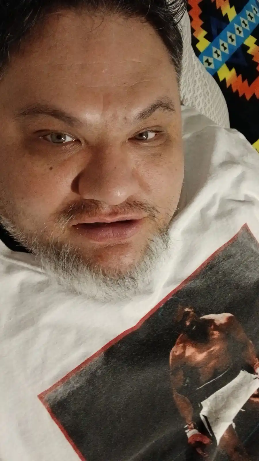 A man with a gray beard and mustache lies in bed wearing a white t-shirt with a graphic of a boxer. He stares directly at…