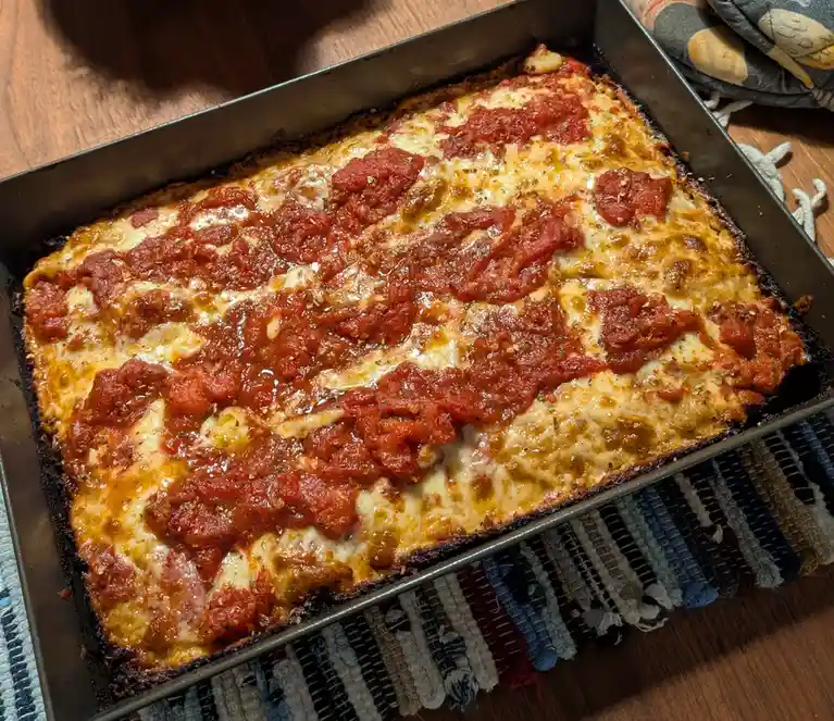 A freshly baked, rectangular dish in a metal baking pan, covered in melted cheese and red tomato sauce, sits on a wooden…