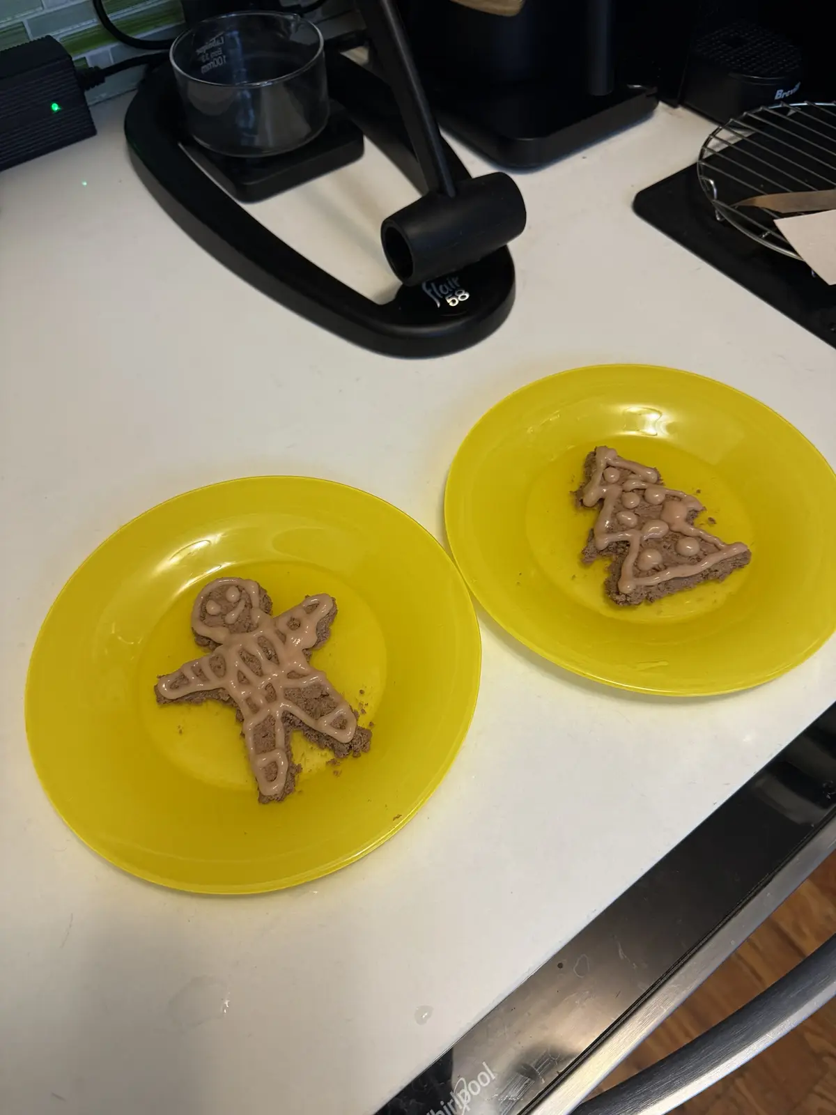 Two yellow plates on a kitchen counter each hold a brown, cookie-like food item decorated with light brown icing: one shaped…