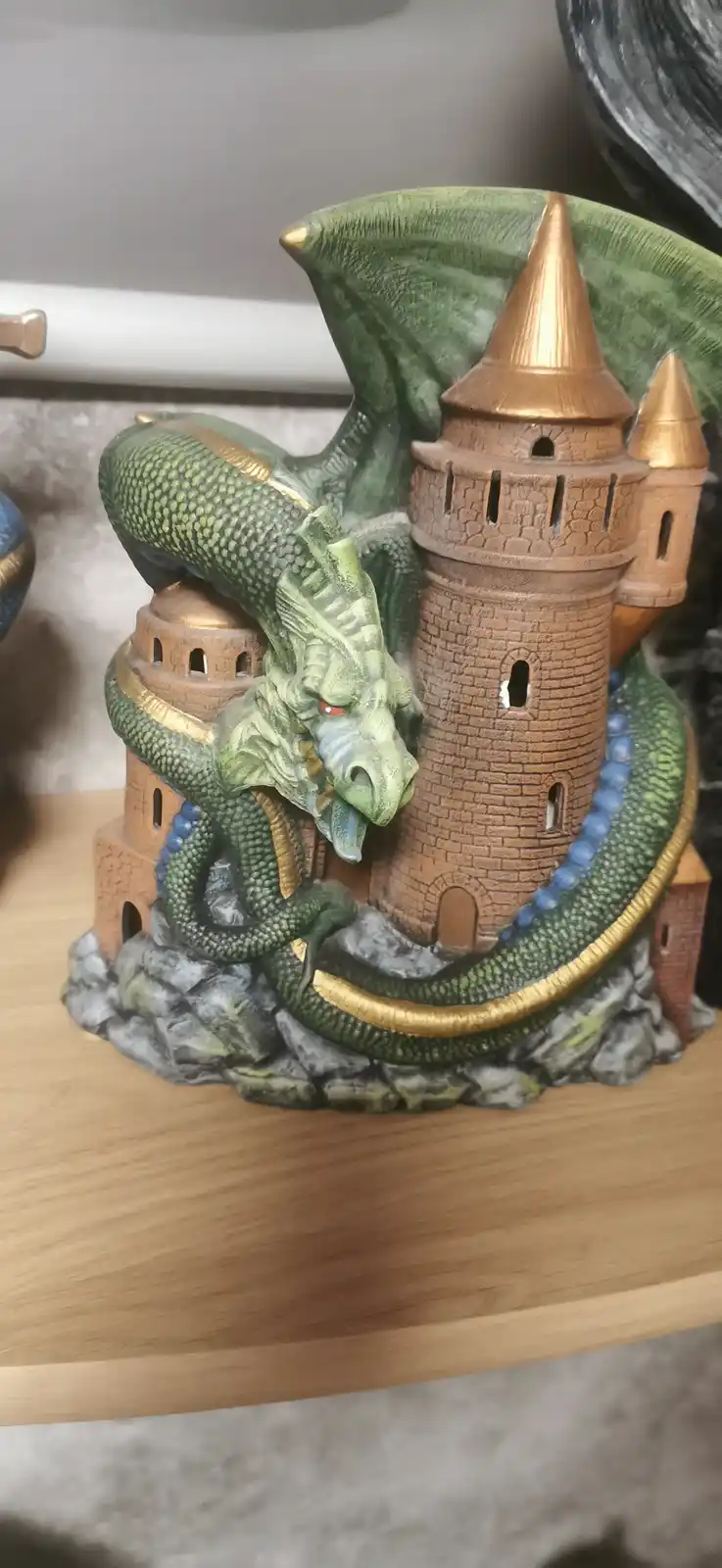 A green dragon figurine with gold accents is coiled around a miniature stone castle with turrets, sitting on a wooden shelf…
