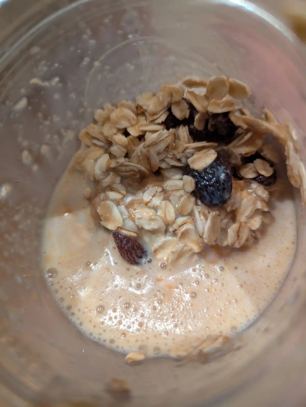 A close-up photo shows a glass bowl containing a mixture of rolled oats, raisins, and a frothy, light-brown liquid…