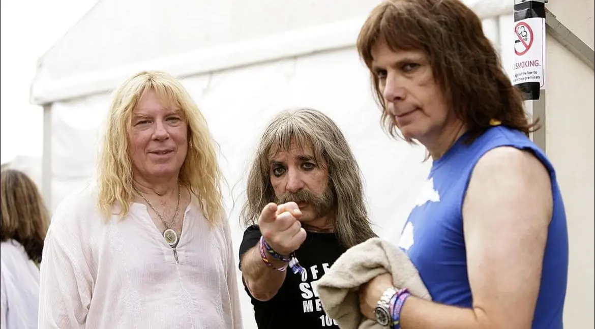 Three men, likely musicians, stand together outdoors near a white tent. The man in the center, with long gray hair and a…