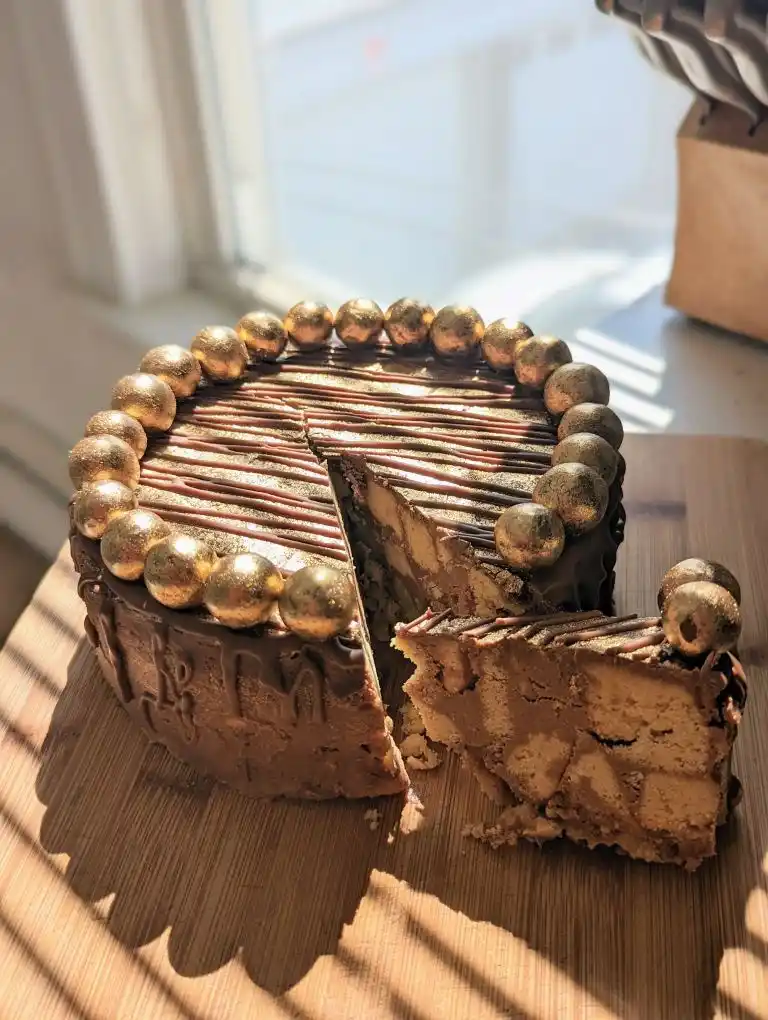 A chocolate cake with gold leaf decorations and a slice removed sits on a wooden board in sunlight. The cake has chocolate…