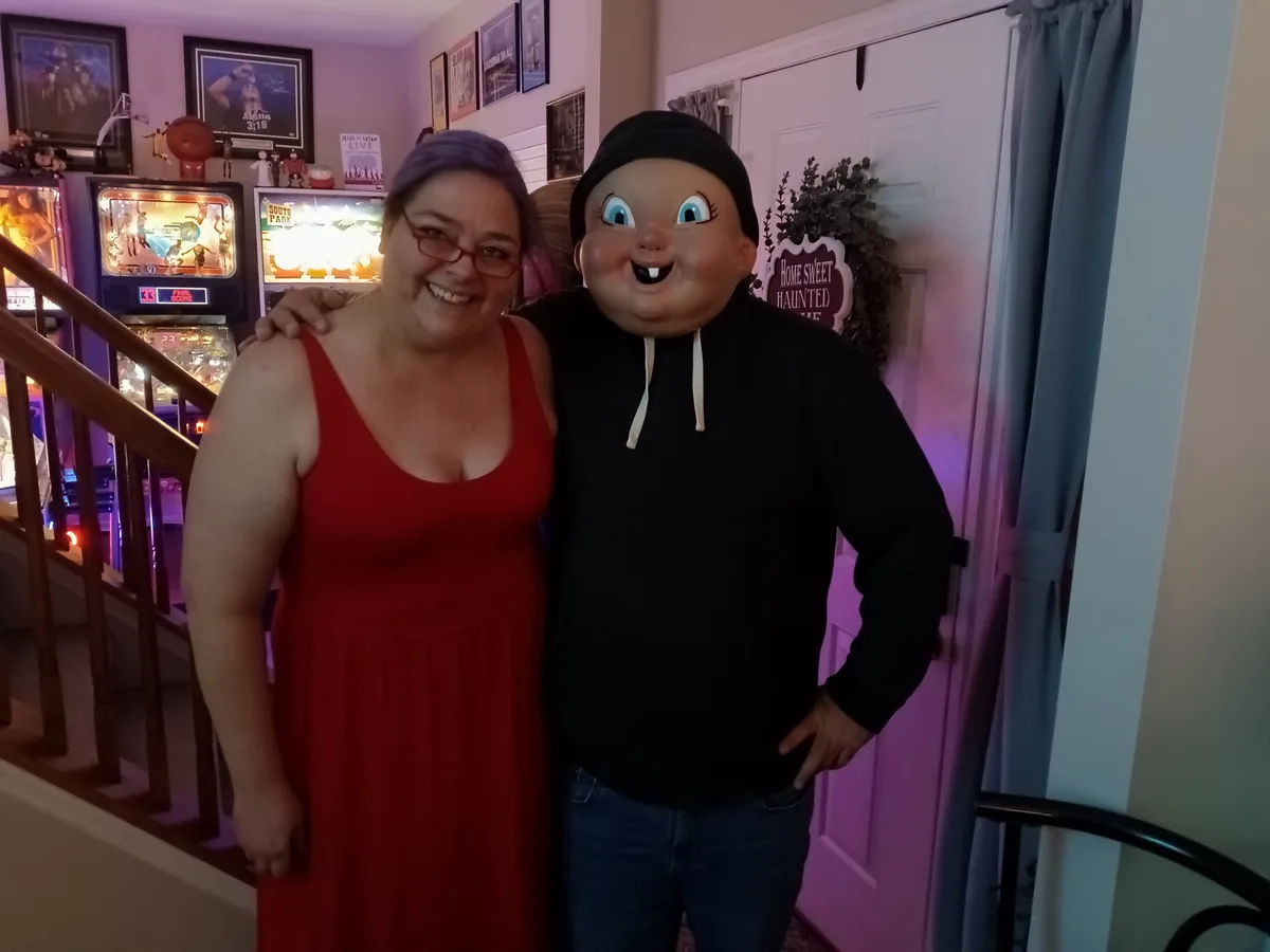 A woman in a red dress smiles beside a man wearing a large, cartoonish, smiling baby-faced mask and a black hoodie. They…