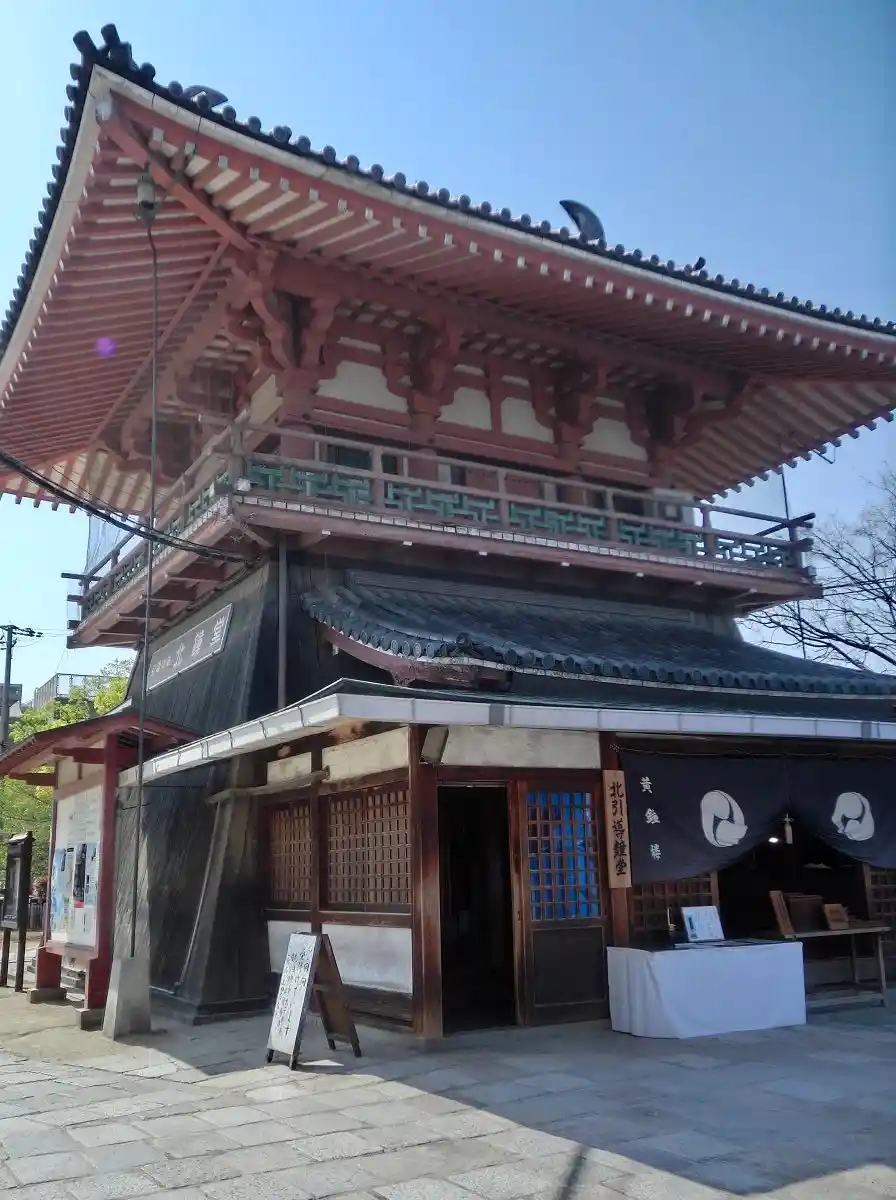 A photo of a traditional Japanese temple building with red wooden beams, tiled roofs, and a stone-paved courtyard. A black…