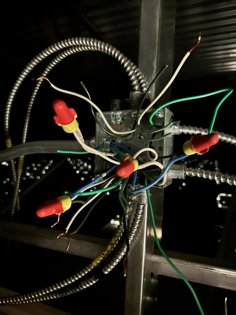 A close-up photo shows a chaotic electrical junction box with numerous wires of different colors (green, blue, white, black)…
