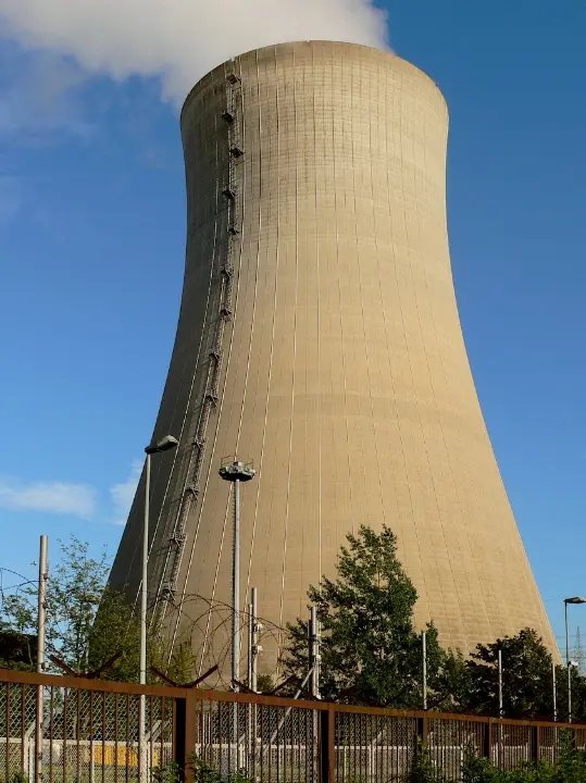 A large, beige nuclear power plant cooling tower stands against a blue sky with scattered clouds. A metal fence with barbed…