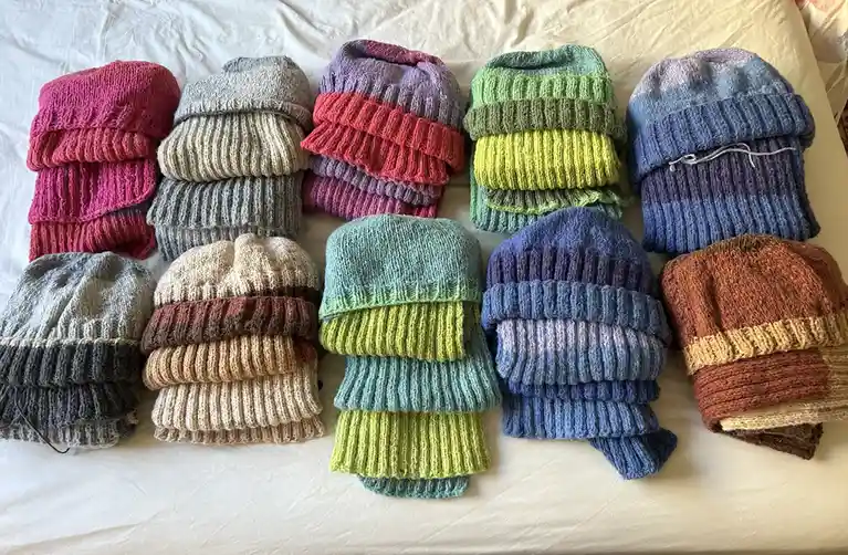 Ten hand-knitted beanies in various colors and patterns are neatly folded and arranged in two rows on a white bedsheet. The…