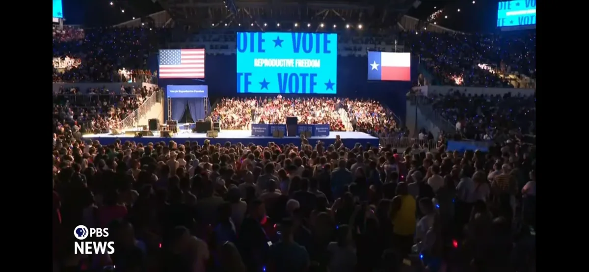 A large indoor rally features a stage with a crowd, flanked by American and Texas flags. A large blue screen displays the…