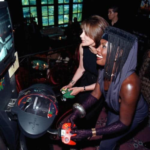 Two women are playing a racing video game on an arcade machine. The woman in the foreground, wearing a purple outfit and a…