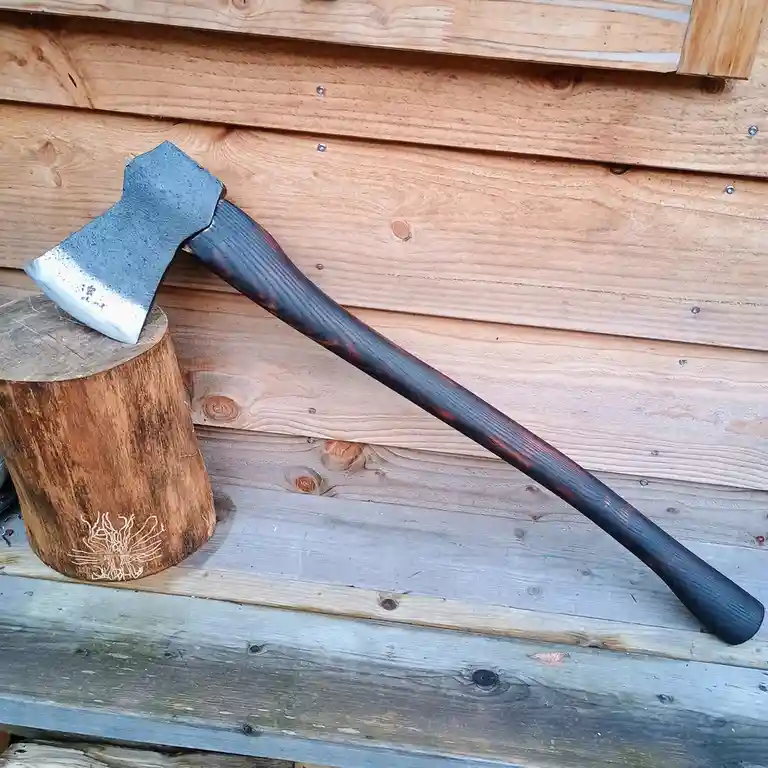 A close-up photo shows a hand-forged axe with a dark, textured wooden handle and a blue-tinged steel head resting on a…