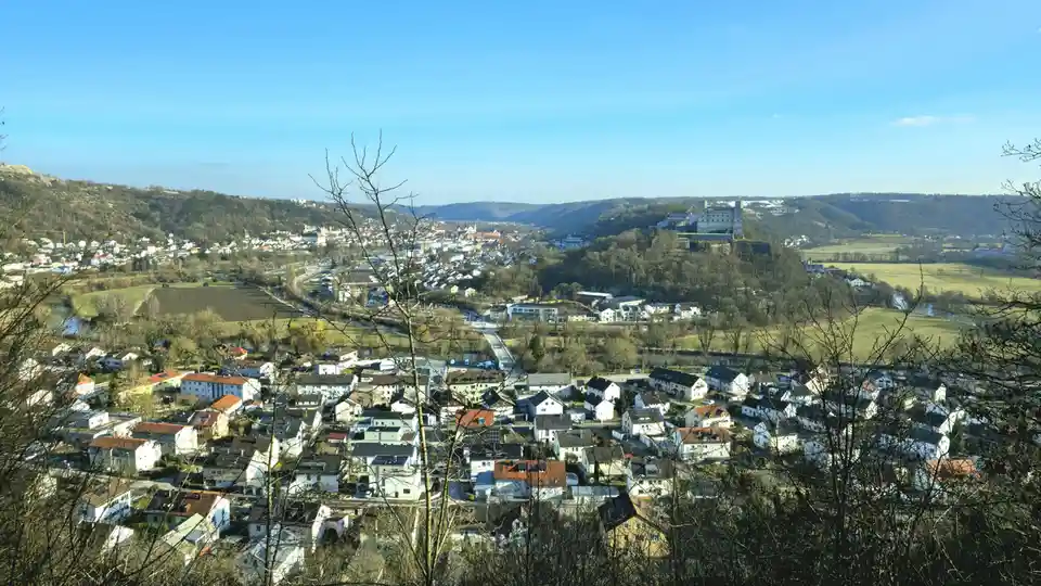 A scenic panoramic view of a European town with houses, a river, and a castle on a hill under a clear blue sky, framed by…