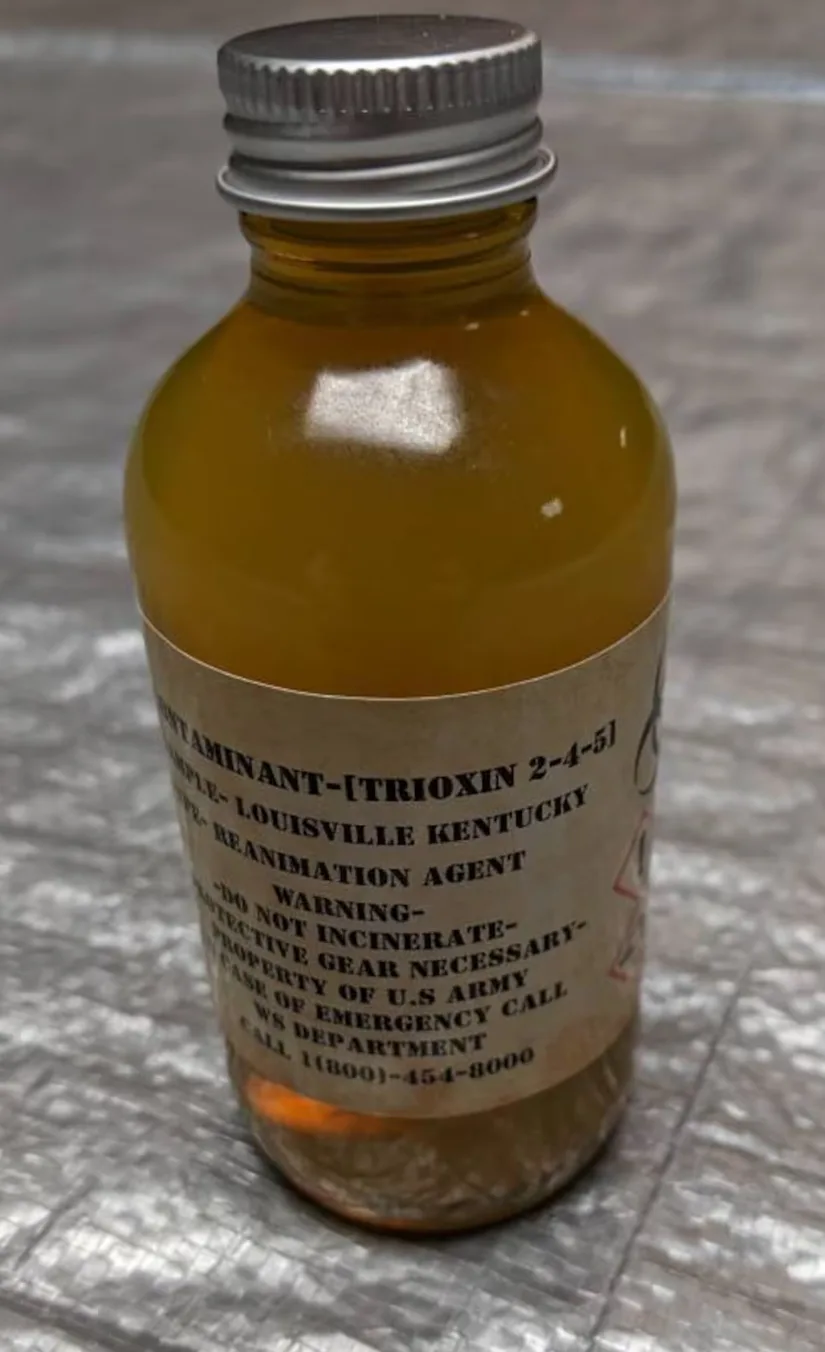 A close-up photo of a small amber glass bottle with a metal cap, sitting on a textured gray surface. The bottle has a beige…