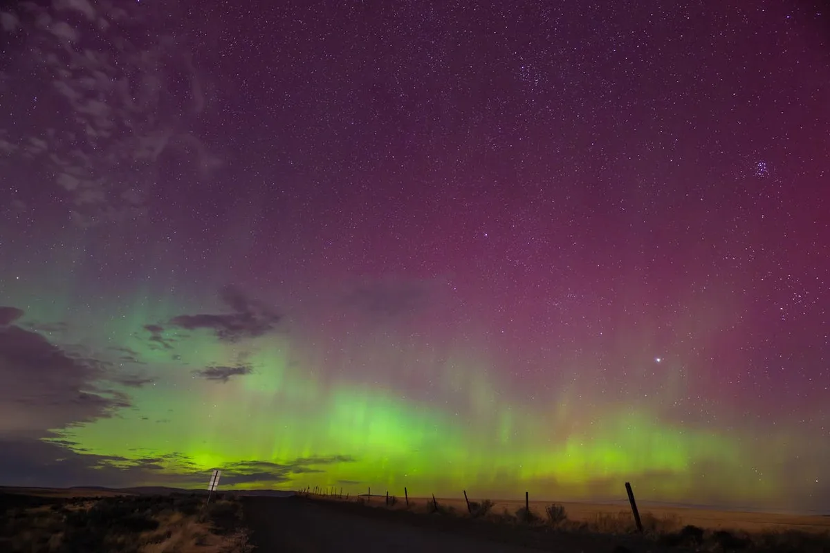 A stunning night sky photograph shows vibrant green auroras dancing across a starry purple and pink heavens above a dark…