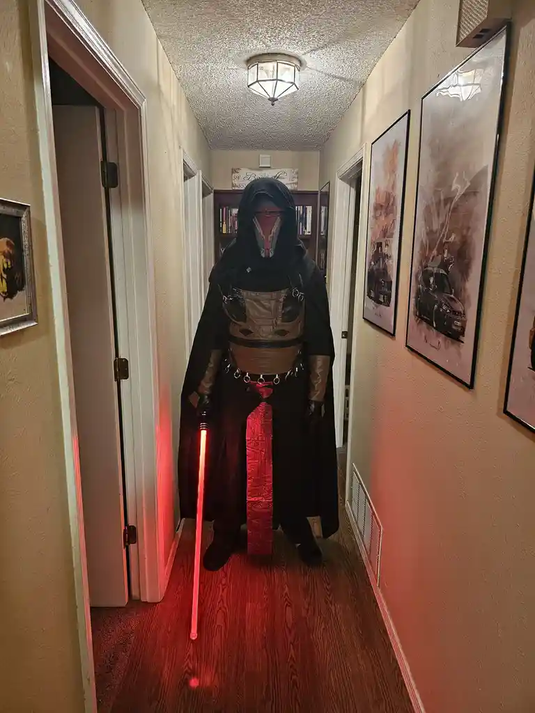 A person in a detailed Star Wars Sith Lord costume, complete with a red lightsaber, stands in a narrow residential hallway…