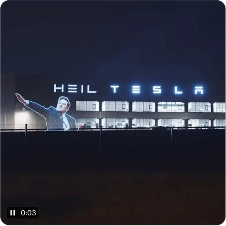 A nighttime photo of a large building with illuminated signage that reads 'HEIL TESLA'. Superimposed over the building is a…
