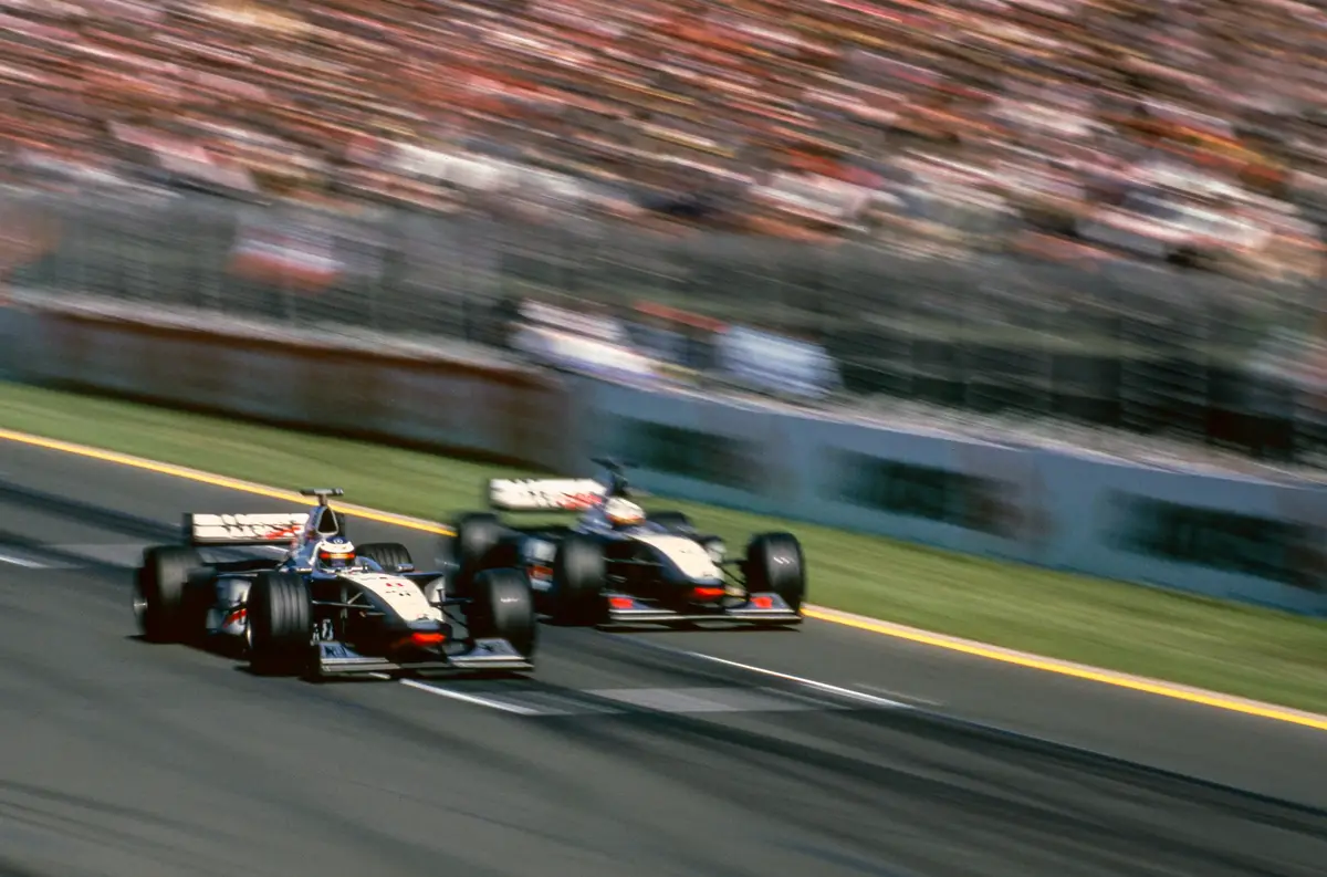 Two Formula 1 race cars are speeding side-by-side on a track, with motion blur conveying high speed. The background shows a…