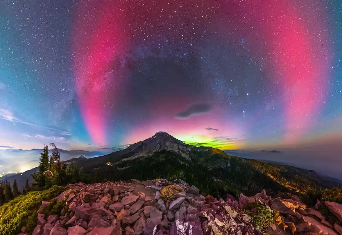 A stunning night sky photograph shows a mountain peak under a vibrant, star-filled sky with pink and green auroras. The…