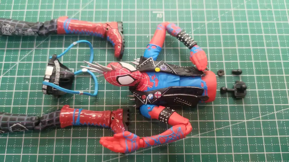A disassembled Spider-Man action figure lies on a green cutting mat, with its limbs and torso separated and scattered…
