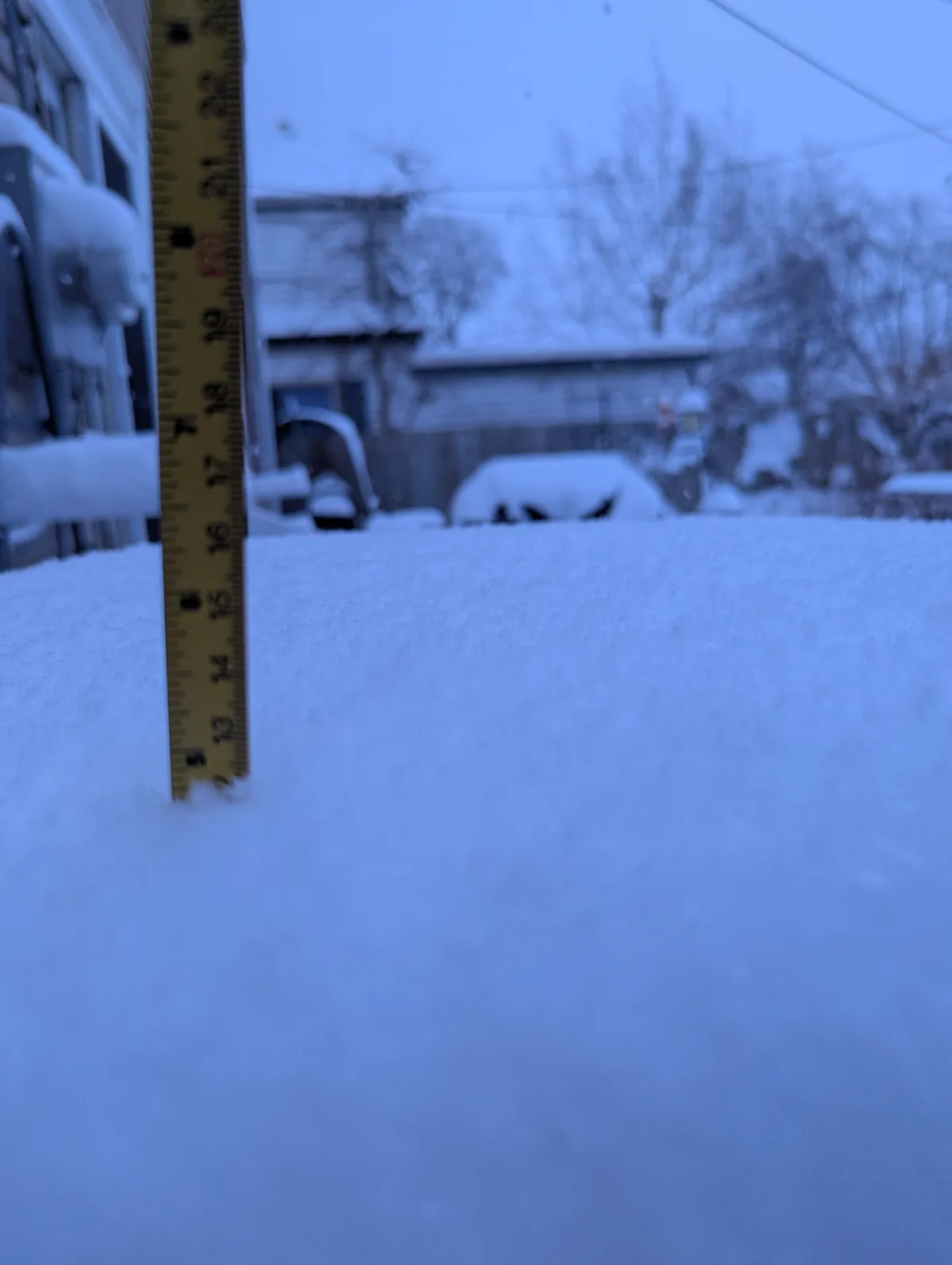 A yellow measuring tape is stuck upright in a thick layer of snow, with the numbers 13 through 21 visible, indicating the…