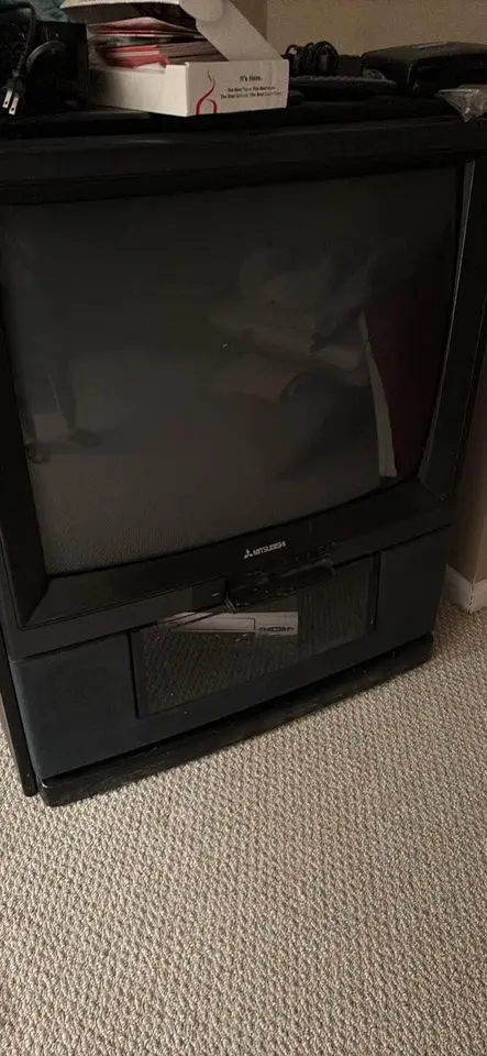 A black Mitsubishi CRT television sits on a beige carpet, its screen dark and reflective, with various items like a tissue…